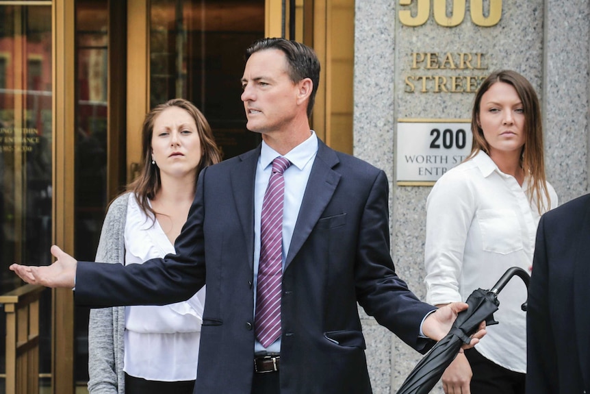 A man in a suit speaks to someone out of the shot, with two women behind him.