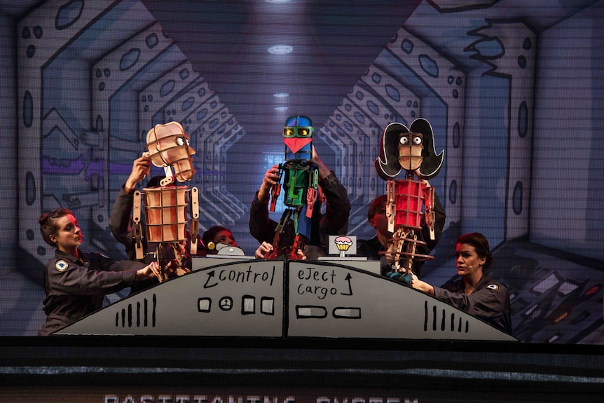 Puppeteers on stage with puppets of Laser Beak Man flanked by two characters, against backdrop drawing of spaceship interior.