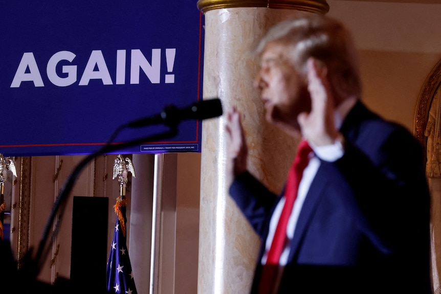 Donald Trump raises his arms as he speaks into a microphone in front of a sign that reads: "AGAIN!"