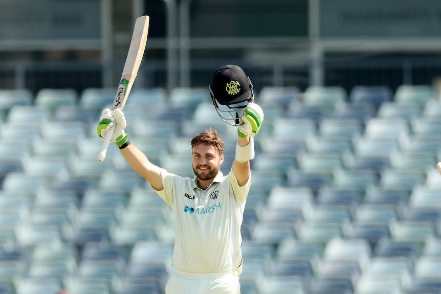 A smiling cricketer holds his bat aloft in one hand and his helmet in the other after completing a hundred in a Shield game. 