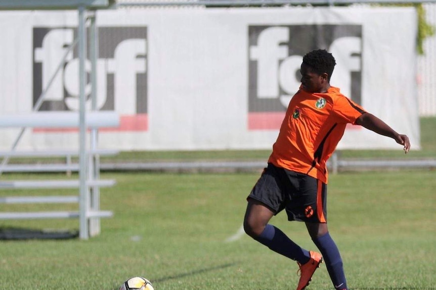 Olivier looks to the ball and kicks it. He wears an orange jersey, black shorts and blue socks.