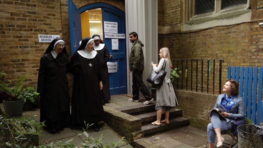 Three people look on while they wait in line to vote as three benedictine nuns leave the polling station at a parish hall.