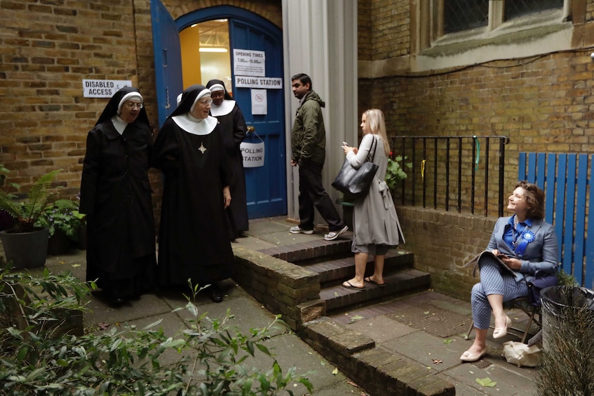 Three people look on while they wait in line to vote as three benedictine nuns leave the polling station at a parish hall.