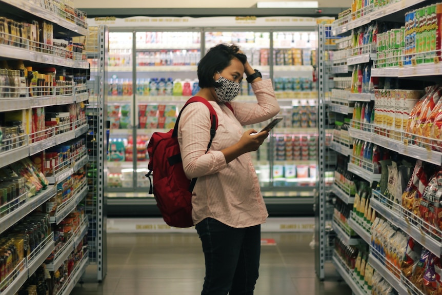 Woman stands in supermarket shopping aisle looking confused as she looks at products.