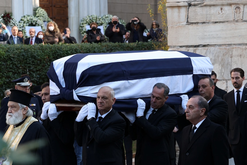 Men wearing black suits are pictured carrying a coffin which has a Greek flag draped around it