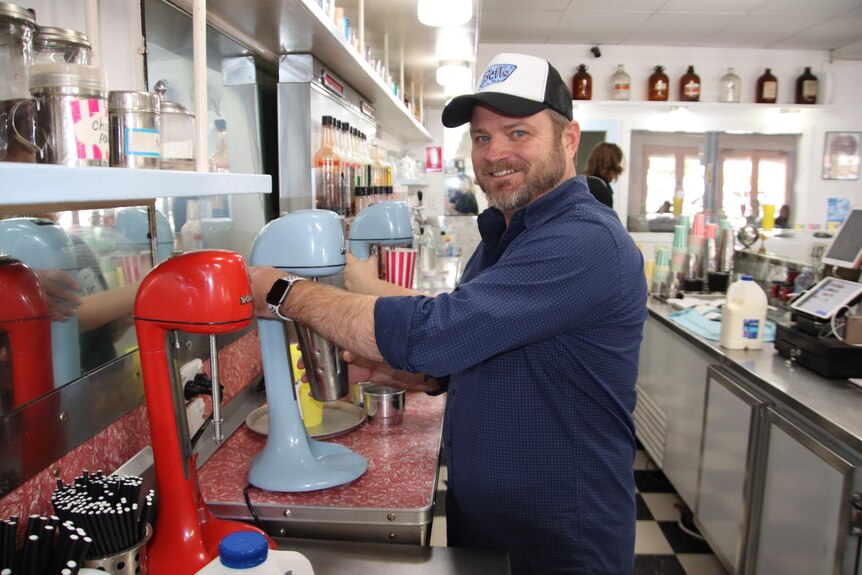 A man wearing a shirt and cap standing next to a milkshake maker holding a cup and smiling