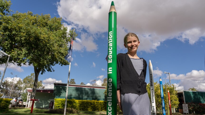 A blonde girl in a stripey dress and cardigan leans against a novelty sized pencil that reads "excellence in education".