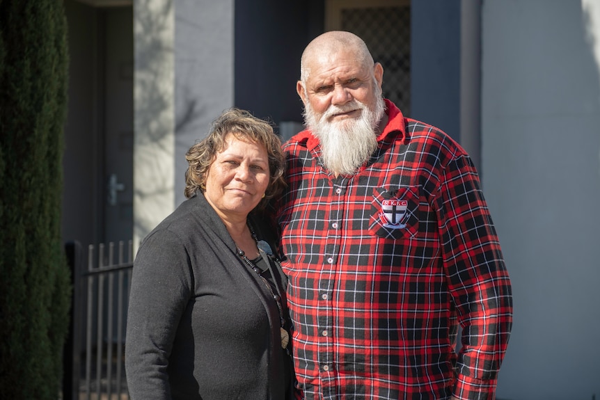 A man and woman stand together outside a house and look at the camera.