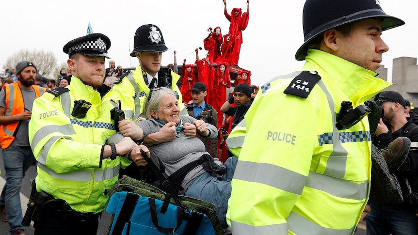 Police officers carry a smiling elderly female protester away from a large crowd