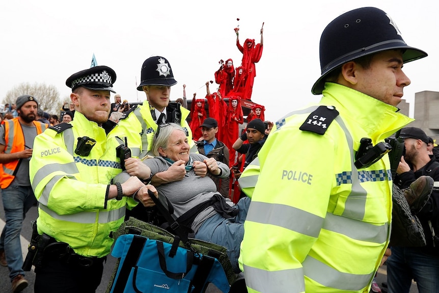 Police officers carry a smiling elderly female protester away from a large crowd