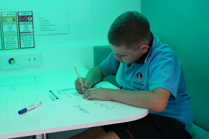 A boy in a blue school polo shirt writes on a worksheet. The lighting is green.