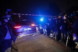 A police car enters the perimeter surrounded by police tape as onlookers and media stand by.