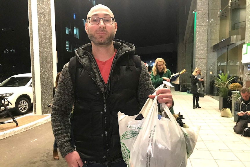 A man carrying a plastic bag full of clothing stands outside a building.