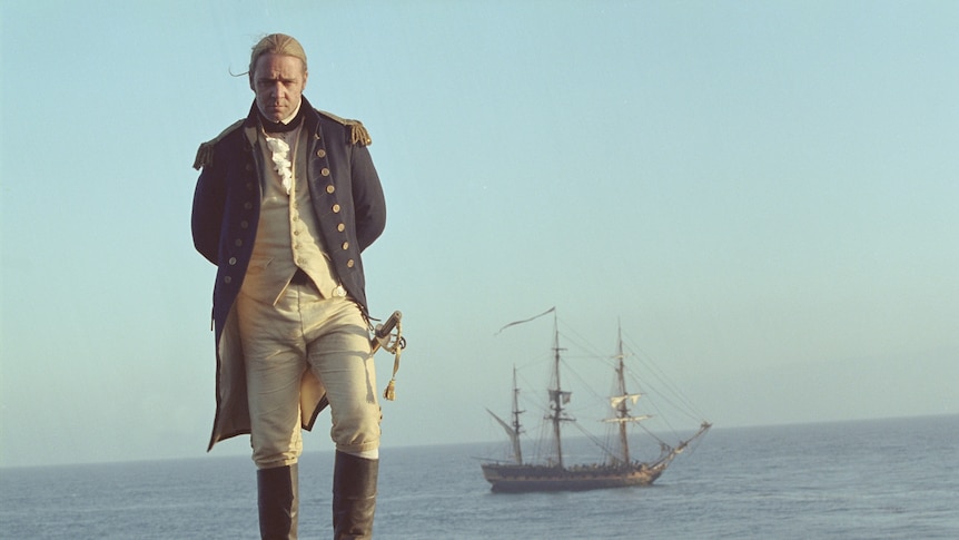 Russell Crowe in an 18th century admiral's uniform standing on a shore with a ship behind