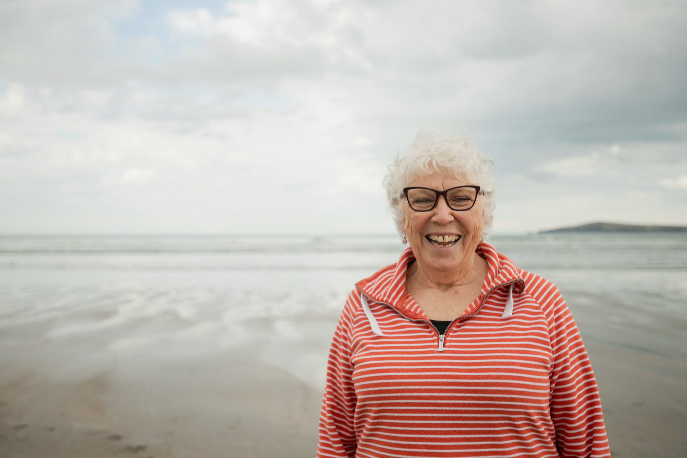 A woman with grey hair and glasses stands on a beach smiling