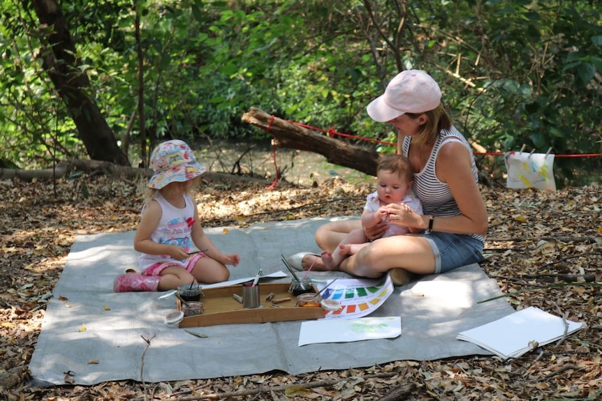 Mum sits on a mat outside with her baby on her lap while her young daughter paints