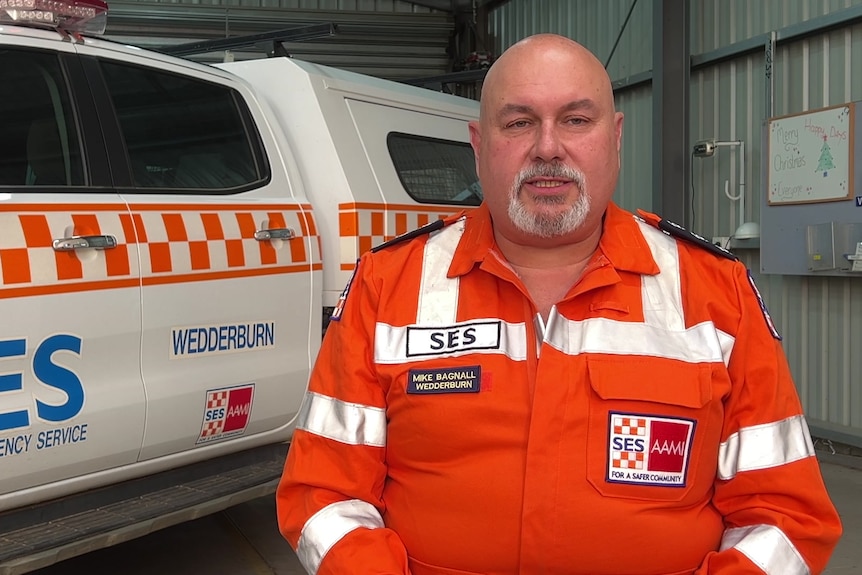A man wearing a bright orange SES uniform, standing in front of an SES vehicle in a garage.