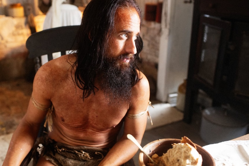 Matt Oxley sits at a kitchen table with a bowl of food, shirtless with long hair and beard.