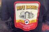 A black t-shirt with an Emu Export beer label on the front with the words Emu Export changed to Wife Basher.