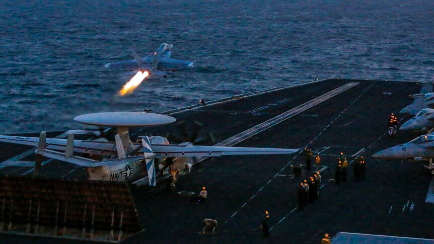 Looking below to the flight deck, a super hornet jet blasts flames from its engines as it takes off from an aircraft carrier.