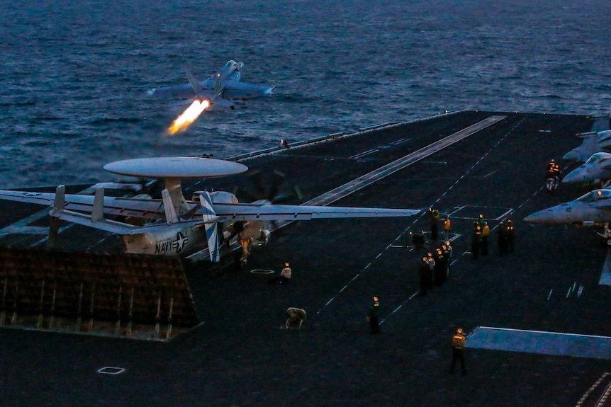 Looking below to the flight deck, a super hornet jet blasts flames from its engines as it takes off from an aircraft carrier.