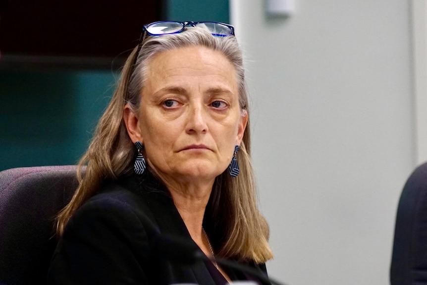 A woman with long, blonde hair and spectacles on her head sits in parliament, looking sombre.