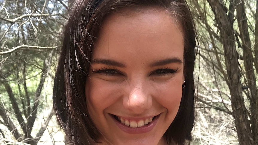 A young woman with brown hair smiles as she poses for a photograph in front of trees.