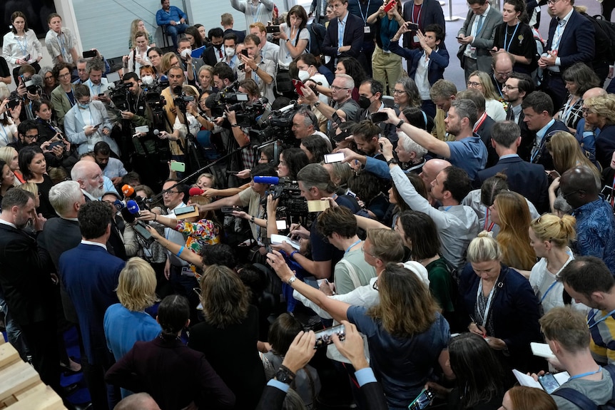 A crowd of media holding out microphones and cameras surround Mr Timmermans. 