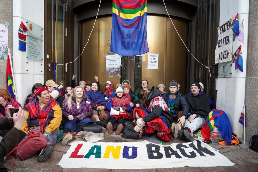 A group of people sit outside a building. A sign laying on the ground in front of them says 'land back'. 
