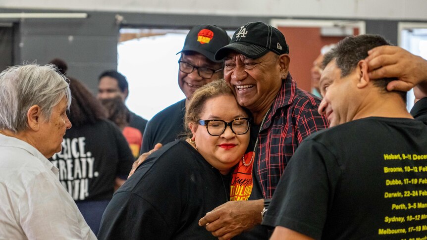 Megan and Nolan hug amid an indoor gathering, with lots of people wearing the colours of the Aboriginal flag and smiling.
