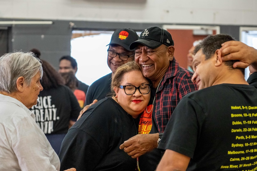 Megan and Nolan hug amid an indoor gathering, with lots of people wearing the colours of the Aboriginal flag and smiling.