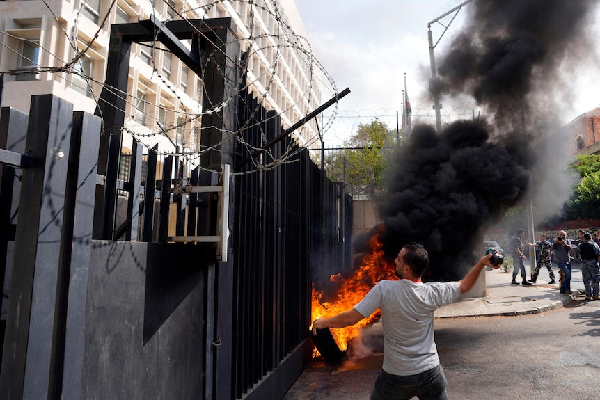 a man in a grey t-shirt throws a molotov cocktail, aiming over a tall black security fence, while a fire burns