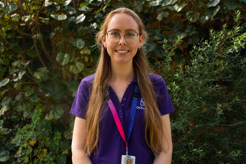 A woman with long hair, spectacles and a purple shirt stands outside posing for a photo.