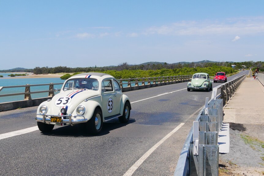 Three Volkswagen Beetles driving on a bridge. There is a lake in the background