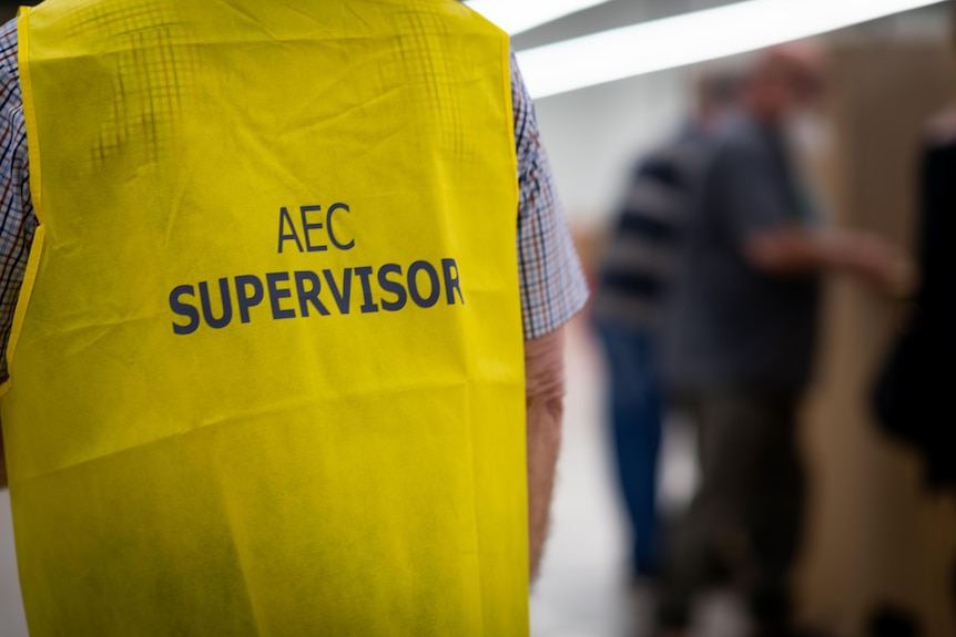 A person wearing a yellow vest reading 'AEC Supervisor', with people voting blurred in the background