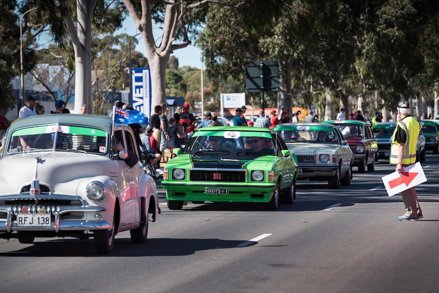 Different kinds of Holden cars, vintage and newer, drive down a street lined with people waving flags and cheering.