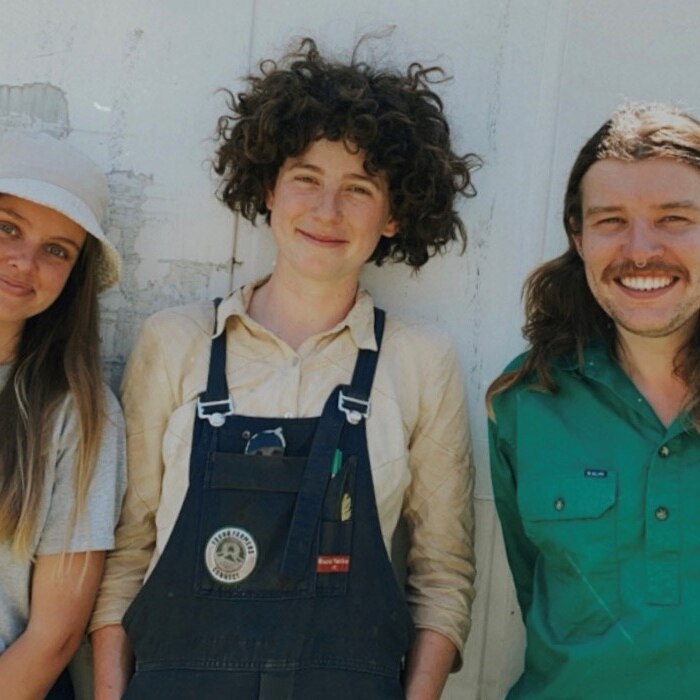 A mid-shot portait of three young white people, wearing gardening clothes, smiling at the camera in front of a white wall