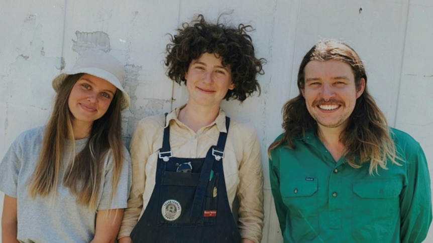 A mid-shot portait of three young white people, wearing gardening clothes, smiling at the camera in front of a white wall