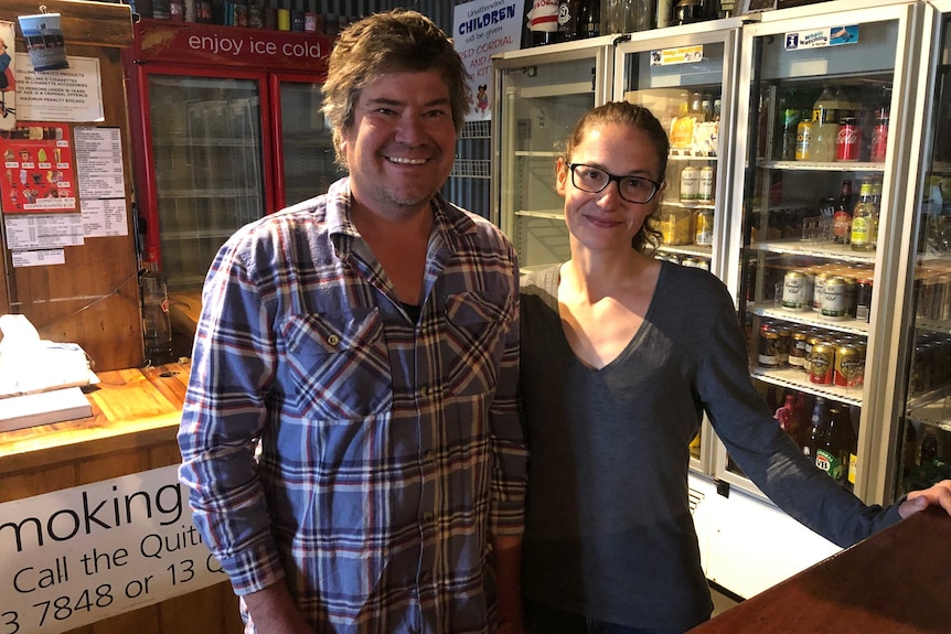A man and a woman stand behind a bar at a pub and smile at the camera