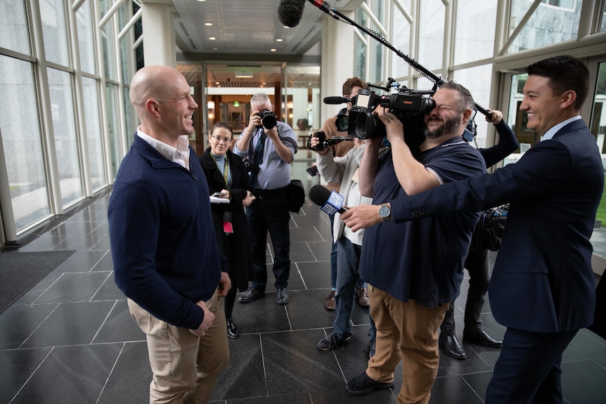 David Pocock smiles with hands in pockets talking with a group of media, some holding cameras, others holding microphones