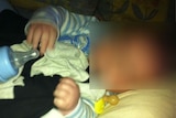 A photograph of a baby with a dummy and a bottle, with his face blurred to prevent identification.