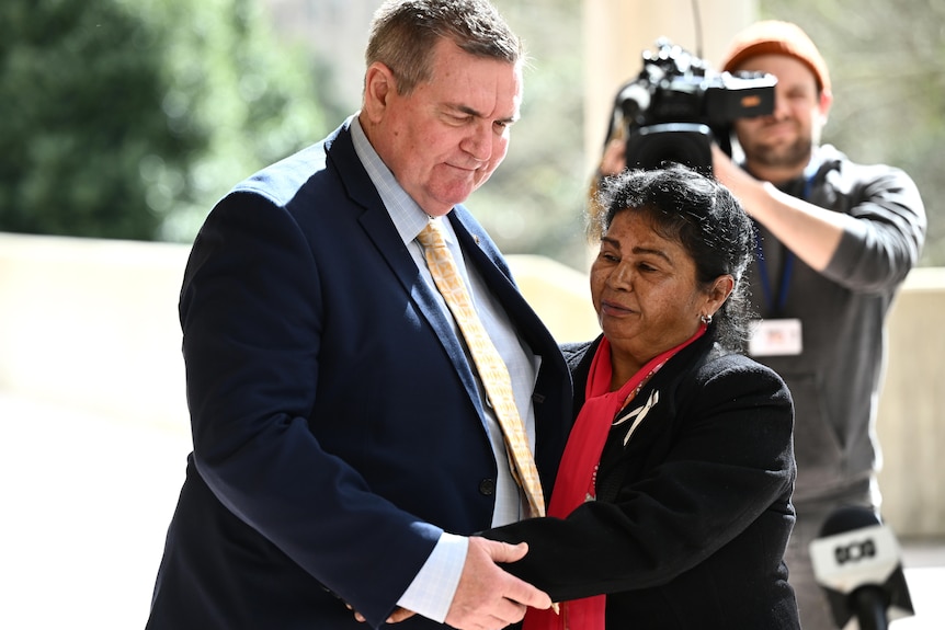 A man and a woman hug with a cameraman in the background. 