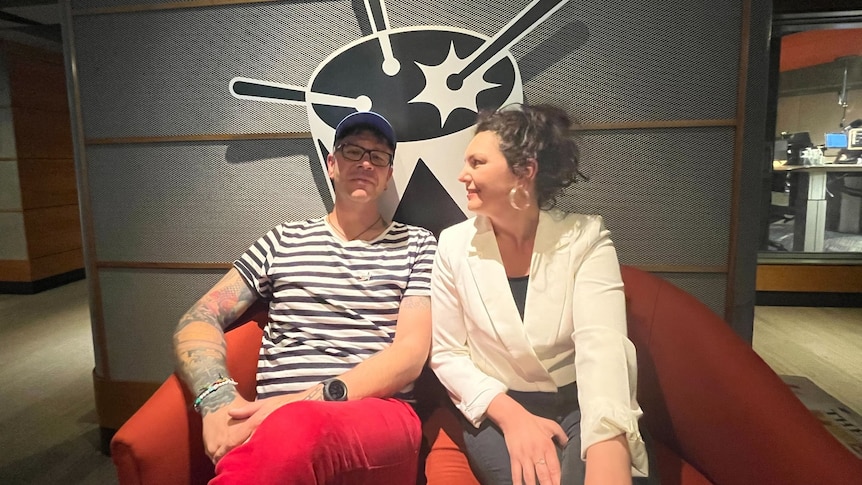 Two people (Dylan Lewis and Eliza Hull) are sitting on a red couch and smiling slightly. A black cardboard drum behind them.