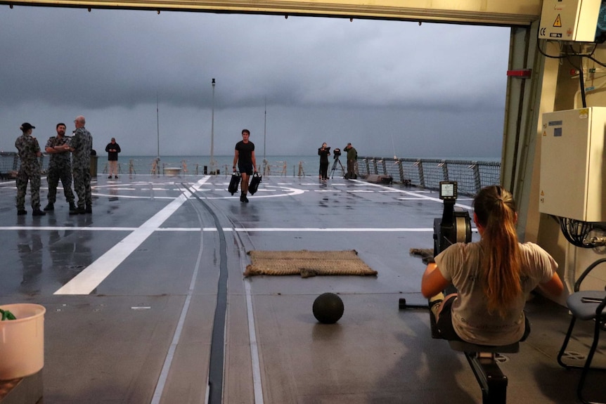 A woman rows on an exercise machine looking out on the deck of HMAS Brisbane.