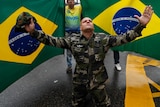 A supporter of President Jair Bolsonaro dressed in fatigues kneels with his arms spread out in front of Brazilian national flag.