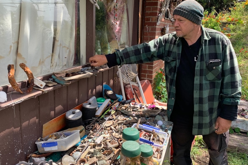 A  man in a beanie looks at his collection of metal items outside his house