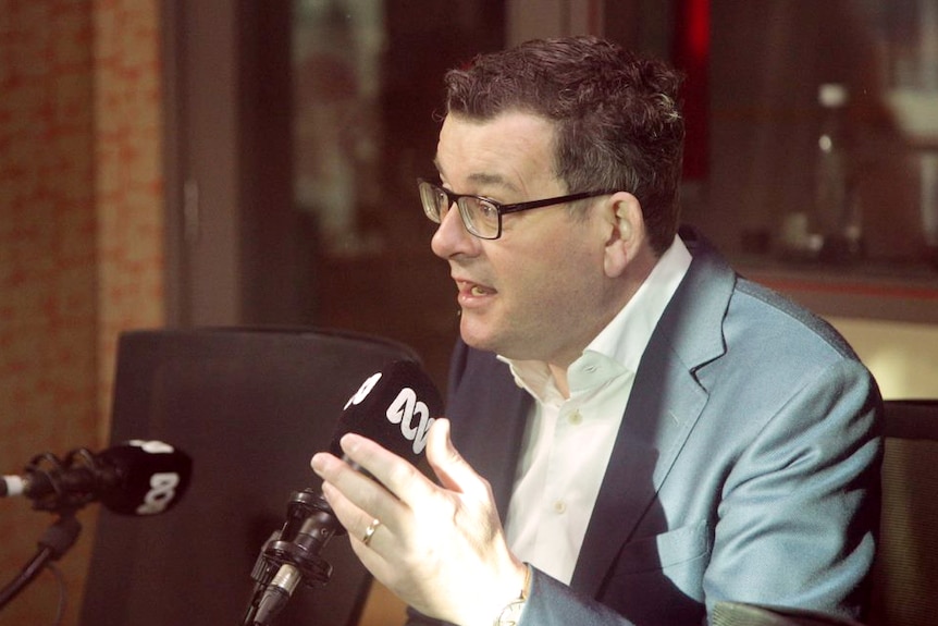 Daniel Andrews, diressed in a suit, sits in front of an ABC-branded microphone.