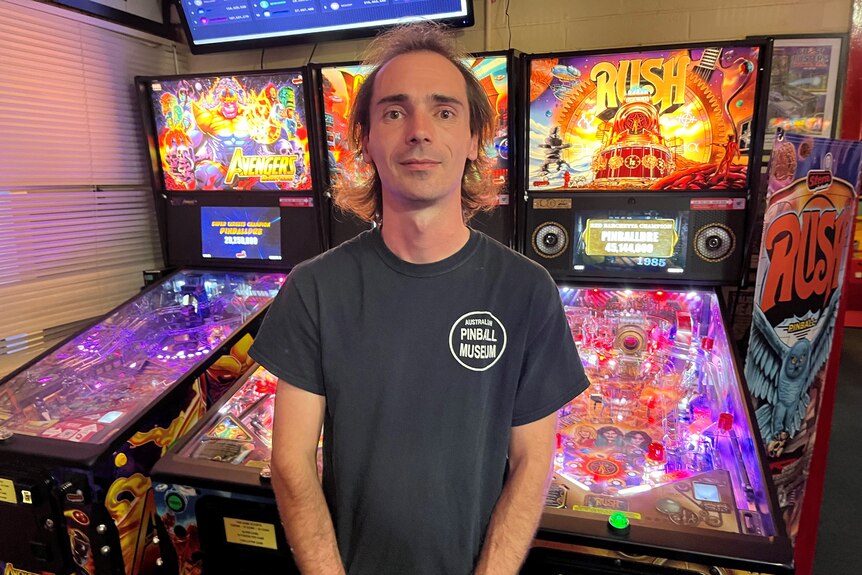 A man with a black tshirt saying pinball museum stands in from of three lighted pinball machines