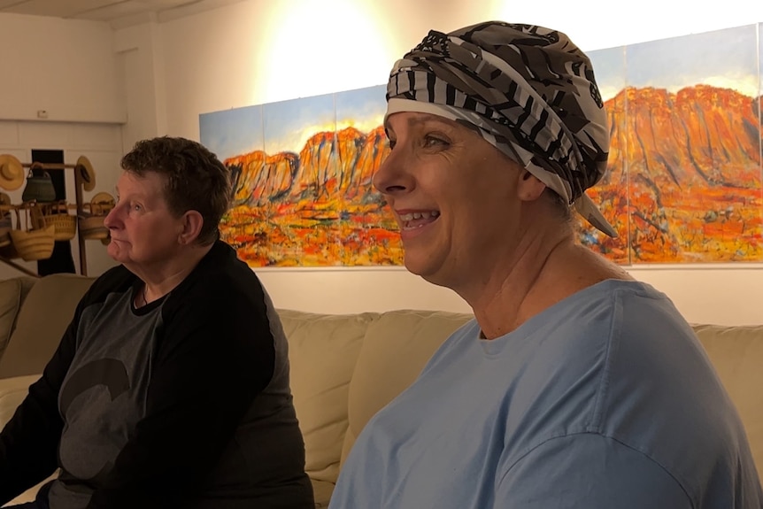 Two women sit on a couch looking away from the camera. One wears a headscarf, a painting of Uluru behind them.