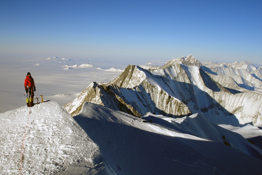 A huge ridge of snow-capped mountains. A person dressed in beanie and orange snow gear stands at the tip of one mountain-top.